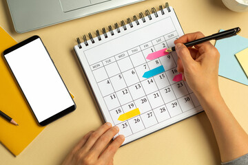 Phone with a white screen and a calendar with dates for writing business meetings. Concept of planning and deadline. A woman takes notes to remind an event in a notebook.