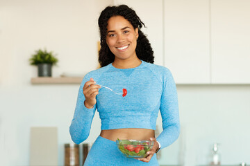 Sport and diet concept. Happy black woman eating fresh salad, standing in kitchen and smiling at camera, copy space