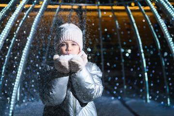 Girl and snow. Girl blowing on palms with snow at night with blue backlight and blurry lights. Holidays theme
