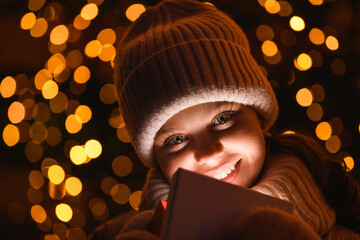 Girl and gift box with Christmas lights. Smiling girl in a hat, scarf looking at the camera at night with hard wrong light, close up. Holidays theme