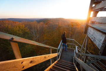 Adventurous young man soaks in the sunrise on stairs of wooden watchtower over autumn foliage trees...