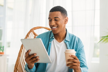 Smiling young black guy in casual chatting on tablet, watch video, enjoy takeaway drink, sitting in...