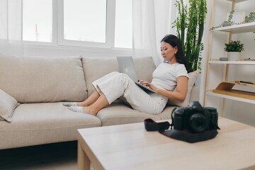 Asian woman photographer lying at home on the couch and reviewing photos from the camera in laptop for color correction retouching, work as a freelance business photographer for yourself home office