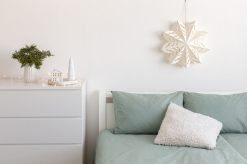 Christmas home decor. Winter bedroom decor. New year interior decorations. White paper snowflake on wall, bed with green linen, fir branch in vase, decorative ceramic house, glowing garland lights.