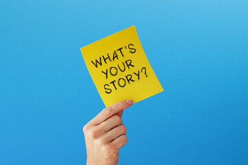 Hand holding note paper with What Is Your Story on blue background