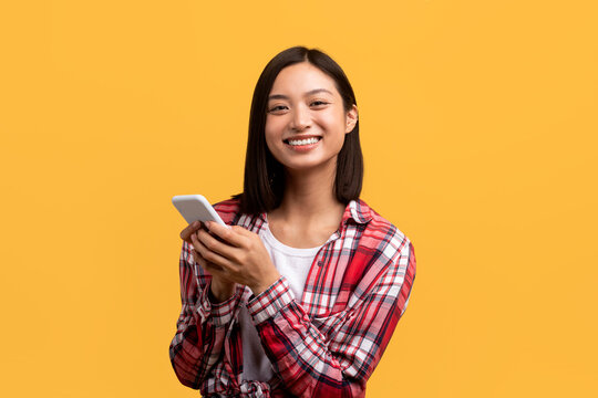 Great app. Excited korean lady with smartphone in hands posing over yellow background, looking and smiling at camera