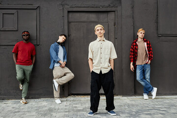 Diverse group of boys wearing street style clothes standing against black wall outdoors and looking...
