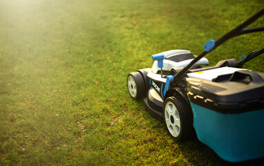 Fototapeta na wymiar A blue lawn mower on the green grass in the park.Mowing grass with an electric mower. Trim the lawn. Cleaning the garden.