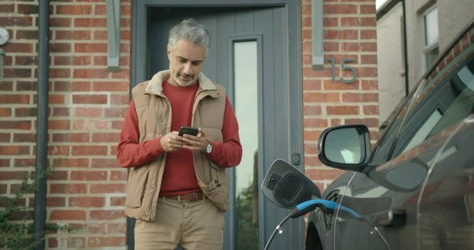 Mid Adult Male Charging Electric Car and using Smartphone app at Home