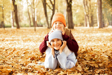 Smiling woman and girl in cute hats lying on each other on ground full of yellow dry leaves. Happy mother and daughter in cozy autumn outfits have fun and walk in golden park. Tree foliage everywhere