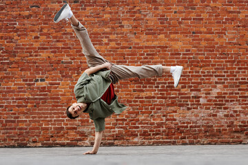 Full length shot of young man doing hip-hop handstand pose and smiling at camera against brick...