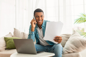 Cheerful young black man in jeans calls by phone, works with documents and laptop, read contract