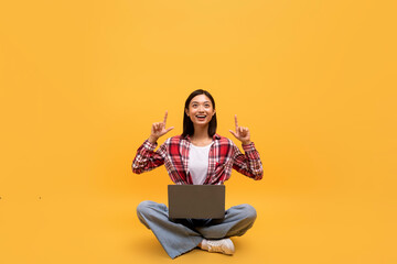 Happy asian lady sitting cross legged with laptop, pointing up at empty space on yellow studio background
