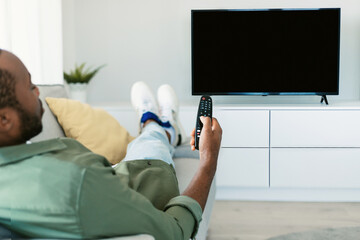 Back view of black man watching TV pointing remote control at empty plasma screen, resting on sofa...
