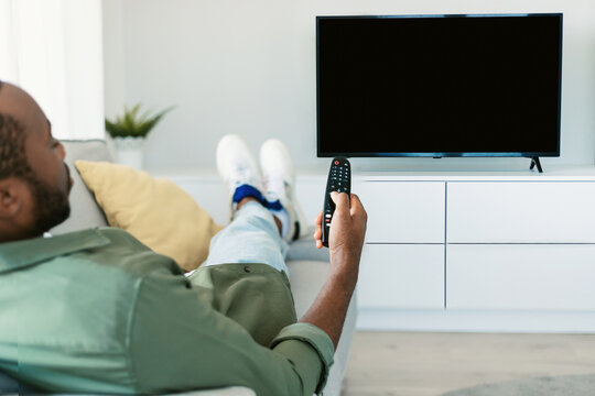 Back view of black man watching TV pointing remote control at empty plasma screen, resting on sofa at home, mockup