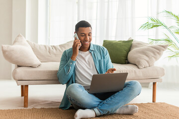 Smiling busy young black male in denim calls by phone, has chat on laptop, sit on floor in living room interior