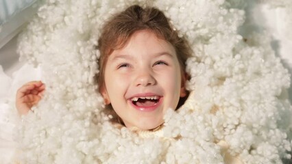 Cheerful little girl joyfully lie in pile of white airly pillow filler and look up closeup, top view, copy space. Only head of kindergarten child on synthetic material background. Active lifestyle