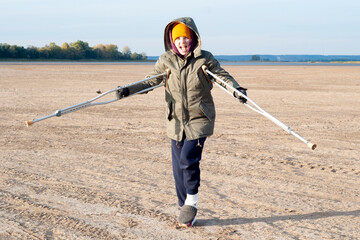 Teenage girl with leg injury and forearm crutches with raised hands and winning gesture, enjoying her vacation on a sunny day, during sunset