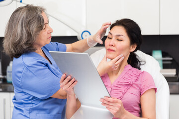 Adult asian woman looking at mirror while experienced female cosmetologist explaining her future hardware facial procedure in medical esthetic office.