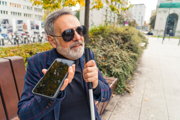 Elegant blind bearded grey-haired mature man with dark sunglasses on holding a walking stick,...