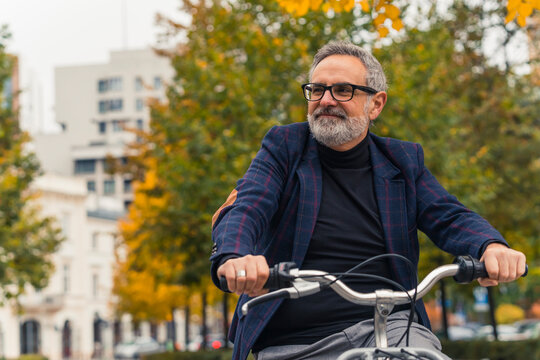 Stylish and happy bearded grey-haired mature man with eyeglasses enjoying warm weather during the fall season while riding a bicycle around trees with yellow leaves. High quality photo