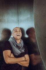 Young man with shaved head, wearing black T shirt, long patterned scarf around neck, crossing arms,...
