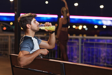 Handsome sporty man drinking lemonade after workout, sitting on a bench on the riverside - 541335267