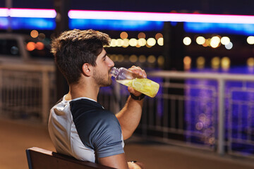 Handsome sporty man drinking lemonade after workout, sitting on a bench on the riverside - obrazy, fototapety, plakaty