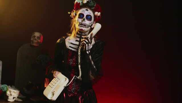 Beautiful woman answering landline phone call, standing over black background with dios de los muertos holiday costume. Celebrating holy mexican halloween tradition with skull body art. Handheld shot.