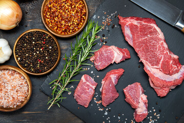 Sliced fresh meat on a black, stone board. Salt, pepper, herbs and spices with vegetables on a dark table. View from above.