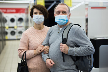 Portrait of European spouses of mature age in protective masks who came to the electronics and home appliance store during ..the pandemic, holding each other by the arms