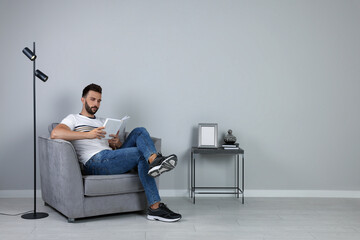 Man reading book in armchair near gray wall, space for text