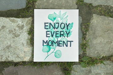 Card with phrase Enjoy Every Moment on ground, top view