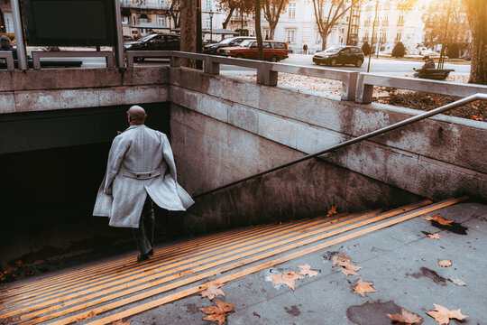 View From Behind Of A Mature Elegant Bald Black Businessman In A Grey Overcoat Descending To A Subway Station, The Floors Of The Coat Flutter In The Wind; A Hairless African Guy Going Down To A Metro