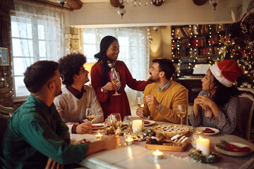 Happy black woman raising toast during Christmas lunch with friends at dining table.