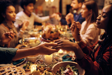 Close up of couple holding hands while praying with friends during Thanksgiving dinner at dining...