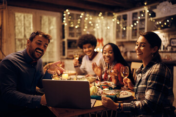 Happy friends making video call via laptop while having dinner at dining table on Thanksgiving.