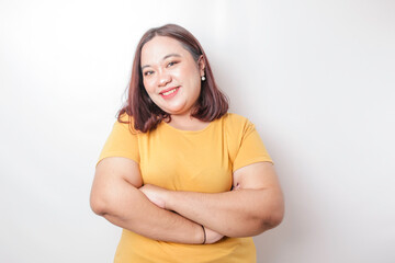 Portrait of a confident smiling big sized Asian woman wearing yellow shirt standing with arms folded and looking at the camera isolated over white background