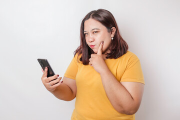 Fototapeta premium The angry and mad face of big sized Asian woman in yellow shirt while holding her phone on isolated white background.