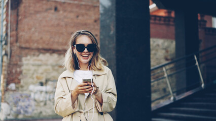 Fototapeta premium Pretty girl is listening to music through earphones using smartphone and dancing walking in the street and enjoying rhythm and modern city. Girl is wearing sunglasses and trendy coat.