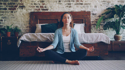 Beautiful young lady is sitting in lotus position on bedroom floor and enjoying meditation, harmony...