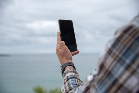Plano detalle de un hombre joven con camisa de cuadros y un reloj inteligente sacando una foto al paisaje  con una mano con su teléfono móvil durante un día nublado en la playa de El Sardinero 