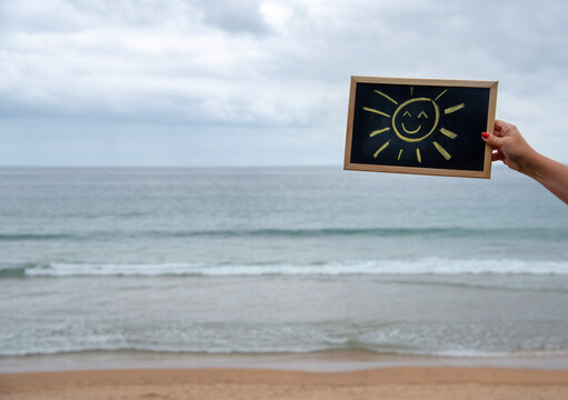 Mano de mujer con uñas rojas sujetando una pizarra negra con bordes de madera con un sol sonriente dibujado con tizas  con una playa de fondo, el mar y las olas durante un día nublado de mal tiempo