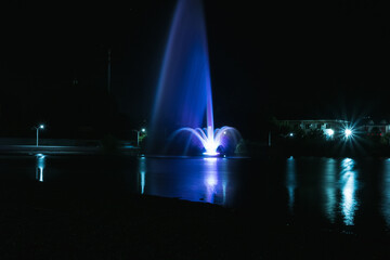 A beautiful high blue with purple fountain in the lake. A night fountain with a reflection in the water. Also in the water are reflected street lights.
