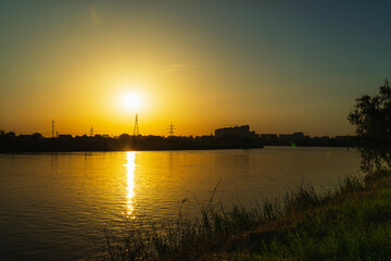 Fototapeta premium Sunset on the background of the river. The orange sun sets over the high-voltage power lines. In the foreground a bank of green grass. Sunset over the river in a small town.