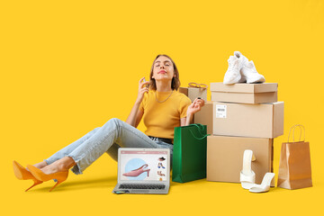 Pretty young woman with new perfume, parcels and laptop sitting on yellow background. Online...