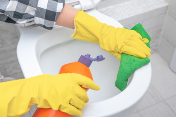 Close up hand with detergent cleaning toilet bowl in bathroom. Man in yellow rubber gloves cleaning toilet seat with green cloth. Bathroom and toilet hygiene. Hand cleaning toilet bowl in bathroom