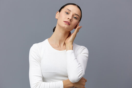 a beautiful, slender, elegant woman stands on a gray background in a white T-shirt with long sleeves and gently touches her neck with her fingers while looking at the camera, smiling pleasantly.