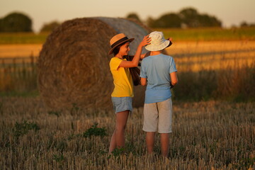 A girl in a straw hat puts a white panama on a boy standing backwards, children stand in a field, a coil of hay behind them, a forest can be seen in the distance.