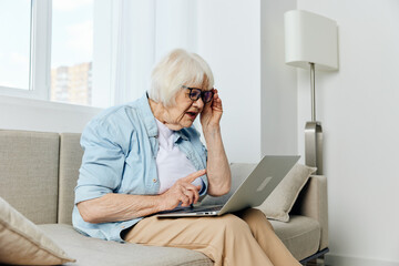 an elderly woman is carefully peering at the laptop monitor trying to read the information holding her glasses with her hand holding it on her lap while sitting on the couch